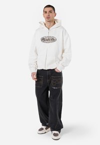 White zip-up hoodie with a logo, paired with dark baggy jeans featuring contrasting stitching and patch pockets, completed with multicolored sneakers.