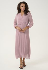 Pleated midi dress in soft pink chiffon, featuring a wrap neckline and long sleeves with gathered cuffs, paired with black strappy sandals.