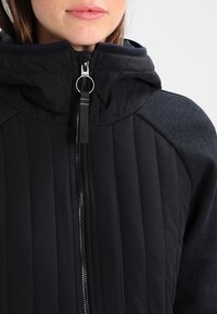 Chaqueta negra aislante con cierre frontal, patrón de acolchado texturizado y acentos en gris oscuro en los hombros. Diseño con capucha y acabado suave.