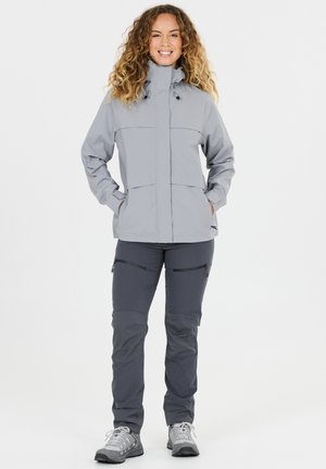 Frau steht, trägt eine hellgraue Kapuzenjacke, dunkelgraue Hosen mit Reißverschlusstaschen und graue Wanderschuhe, lächelt mit den Händen in den Taschen.