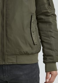 Olive green padded bomber jacket with a zip closure, ribbed cuffs, and hem, featuring side and arm pockets for storage.