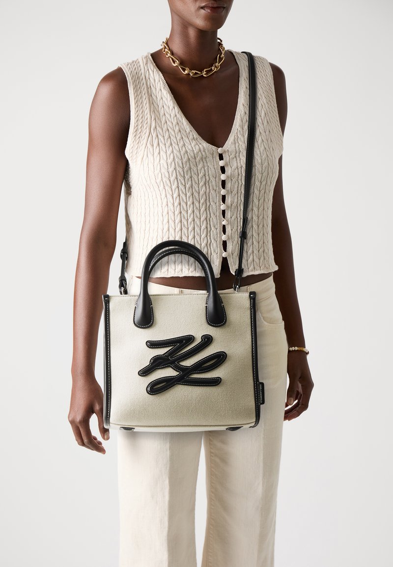 Sac à main en toile beige avec des accents en cuir noir, présentant un design brodé. Poignées courtes et une bandoulière amovible sont incluses.