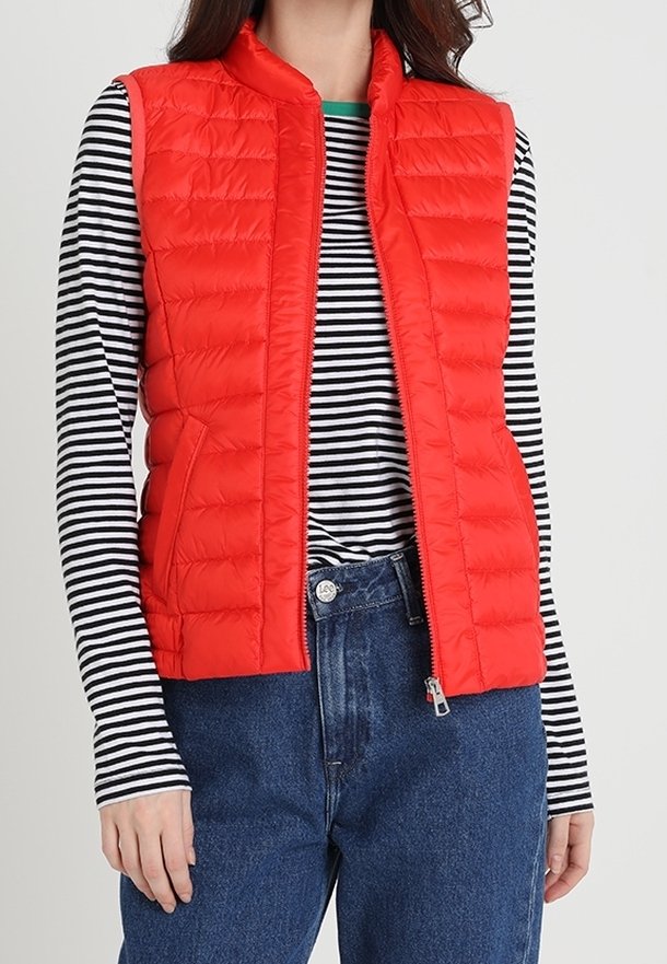 Röd vadderad väst med ståkrage och dragkedja framtill, bärs över en svart och vit randig långärmad tröja. Jeans fulländar outfiten.