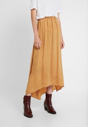 Femme portant une jupe midi asymétrique couleur moutarde avec taille élastique, des bottines marron foncé texturées, haut blanc à manches courtes.