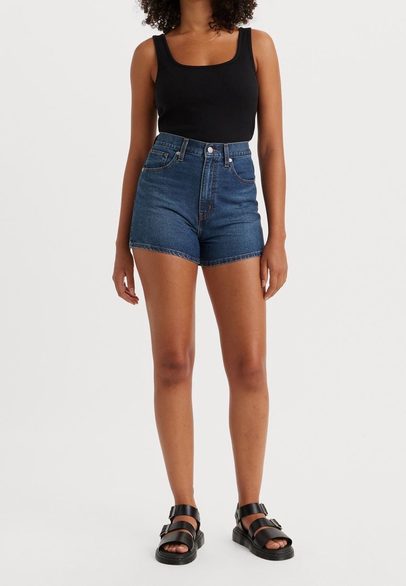 Shorts en denim bleu foncé, taille haute avec un ourlet brut. Associés à un débardeur noir et des sandales à talons noires. Texture lisse.