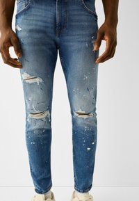 Modré džíny z denimu s přiléhavým střihem, které mají popraskané oblasti a skvrny od barvy. Zahrnují roztrženiny na kolenou a mírné blednutí.