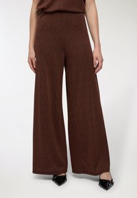 Pantaloni larghi marroni con un motivo geometrico, realizzati in tessuto morbido e con una superficie liscia, abbinati a scarpe nere appuntite.