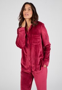 Conjunto de loungewear de terciopelo en un profundo color rojo, que incluye una camisa de botones con un bolsillo en el pecho y pantalones a juego. Textura suave y corte relajado.