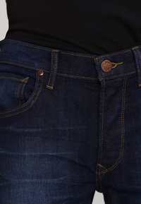 Jeans de mezclilla azul oscuro con costuras amarillas, cierre con botón y marca "Lee", bolsillos delanteros y traseros, y una textura suave.