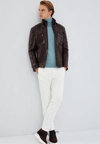 Giacca di pelle marrone con colletto alto, dolcevita azzurra chiara, pantaloni bianchi e scarpe da ginnastica marrone scuro con accenti testurizzati.