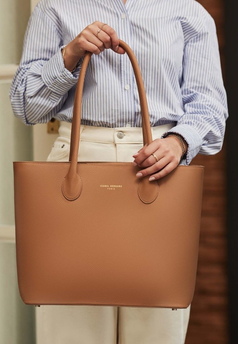 Braune Ledertasche mit verlängerten Griffen, ausgestattet mit einer strukturierten Oberfläche und einem goldenen Logo. Einfaches Design mit geräumigem Innenraum.