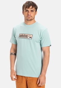 Světle tyrkysové bavlněné tričko s logem Quiksilver v černé a béžové barvě, kulatým výstřihem, krátkými rukávy a volným střihem.