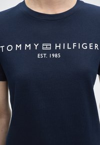 Μπλε ναυτικό μπλουζάκι με λευκό λογότυπο Tommy Hilfiger και την επιγραφή "EST. 1985" στο στήθος, που το φορά ένα άτομο.