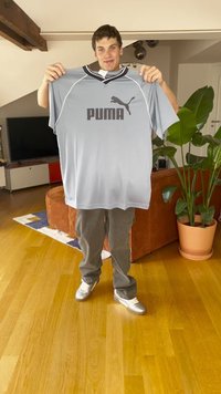 Svetlo siva Puma športna majica s kratkimi rokavi, črn ovratnik in črn logo. Model drži majico pred modernim notranjim okoljem.