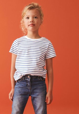 Jong kind met krullend blond haar, gekleed in een wit-blauw gestreepte blouse en blauwe spijkerbroek, staand tegen een oranje achtergrond.