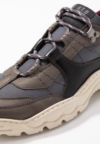 Šedá a černá turistická bota s vrchní částí z polyesteru a kůže, která má šňůrky, polstrovaný límec a strukturovanou podešev s výrazným vzorem.
