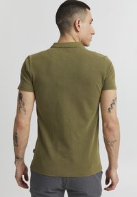 Olijfgroene polo met korte mouwen, geribbelde textuur en een klassieke kraag. Bevat tatoeages op de linkerarm en een genaaid label aan de onderzijde.