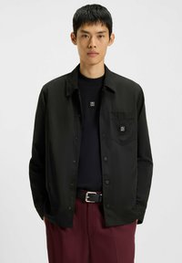 Sort button-up jakke med en glat tekstur, udstyret med en brystlomme og logo detalje. Bæret over en sort sweater og maroon bukser.