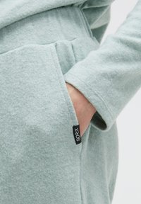 Hellgrau-grünes, weiches Fleece-Kleidungsstück mit einer Tasche; verfügt über ein Etikett mit der Aufschrift "socx." Nahaufnahme des strukturierten Stoffes und des Ärmel-Designs.