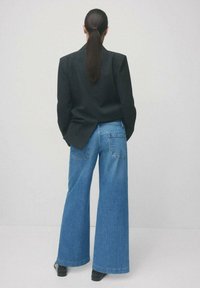 Czarna marynarka, niebieskie jeansy o szerokich nogawkach z dwoma tylnymi kieszeniami oraz czarne botki za kostkę. Gładka tekstura i luźny krój. Gładkie tło.