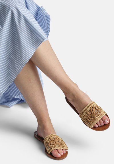 Pieds portant des sandales tissées marron clair à enfiler avec des motifs décoratifs, associées à une jupe ou une robe rayée bleue et blanche.