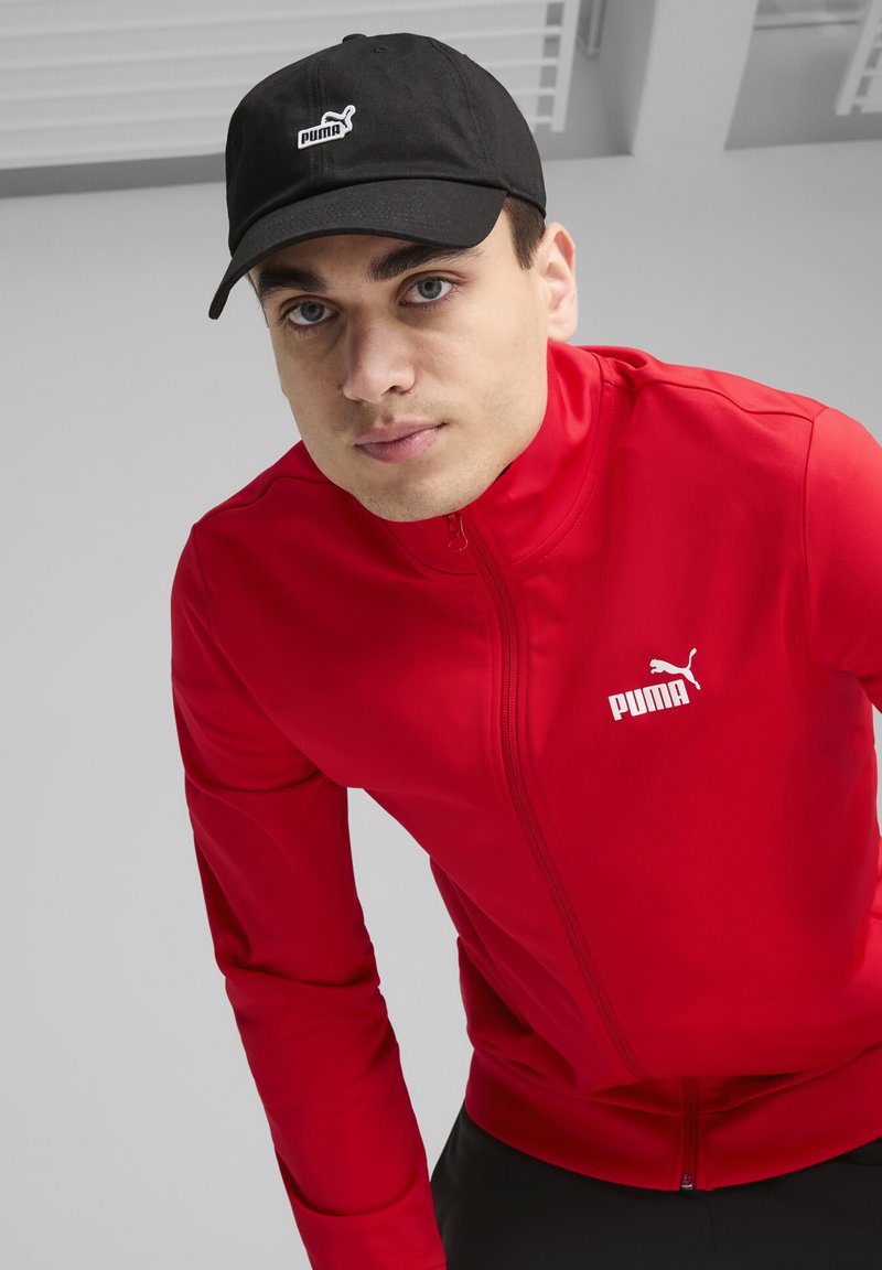 Veste zippée rouge avec un logo Puma, associée à une casquette de baseball noire arborant un logo Puma blanc. Tissu lisse, coupe athlétique, design minimaliste.