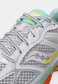 Κοντινή λήψη ενός λευκού αθλητικού παπουτσιού από δίχτυ με κορδόνια με μαύρες ρίγες, γλώσσα σε τυρκουάζ χρώμα, κίτρινες λεπτομέρειες και σόλα σε πορτοκαλί και πράσινο χρώμα.