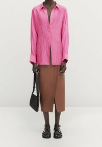 Roze overhemd met knopen en een losse pasvorm, lange mouwen, gecombineerd met een bruine rok tot de knie met een split aan de voorkant, aangevuld met sandalen met bandjes en een geweven tas.