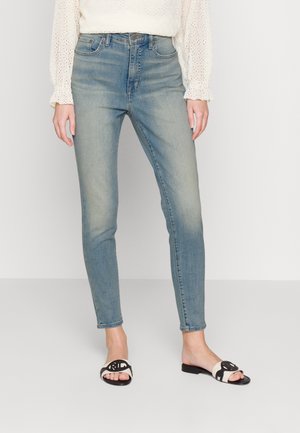 Mujer con vaqueros pitillo azul claro, blusa blanca de manga larga y sandalias abiertas en blanco y negro, de pie sobre un fondo liso.