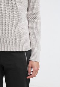 Hellgrauer gestrickter Pulloverärmel und -bündchen am linken Arm neben einer Hand, die an einer schwarzen Hose mit sichtbarem silbernem Reißverschluss liegt.