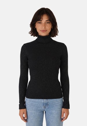 Donna con capelli corti e scuri che indossa un maglione nero a coste con collo alto e jeans azzurri, in piedi davanti a uno sfondo chiaro uniforme.