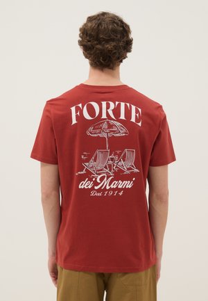 Persona con cabello rizado que lleva una camiseta roja con estampado blanco de sillas de playa, paraguas, bebidas y el texto "FORTE dei Marmi Dal 1914" en la espalda.