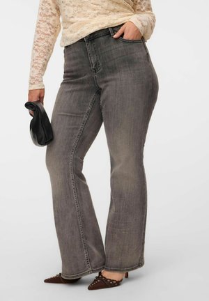 Personne portant un jean gris délavé taille haute avec jambes évasées, un haut en dentelle crème à manches longues, des chaussures pointues marron cloutées, tenant une pochette noire.