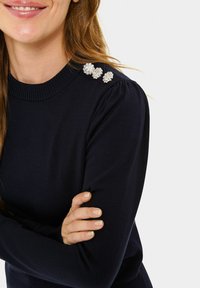 Granatowy sweter z dzianiny z prążkowanym okrągłym dekoltem, z trzema ozdobnymi dodatkami w kolorze srebrnym na lewym ramieniu.