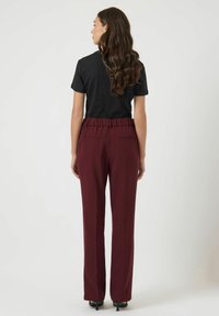 Kobieta z długimi falowanymi włosami, ubrana w czarną bluzkę z krótkim rękawem i wysokim stanem bordowe spodnie, stojąca tyłem na jednolitym tle.