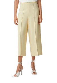 Helle gelbe Culotte-Hosen mit vorne gelegten Falten, hoher Taille und weiter Beinform. Kombiniert mit weißen High-Heelsandalen mit Knöchelriemen.
