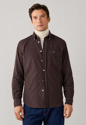Hombre con camisa marrón oscuro abotonada sobre un jersey de cuello alto blanco y vaqueros azules contra un fondo claro y liso.