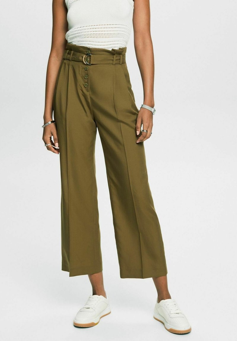 Personne portant un pantalon ample vert olive avec ceinture et boutons, un haut en maille blanc sans manches, des baskets blanches, des bracelets et une montre.