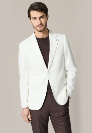 Mężczyzna ubrany w biały blezer na ciemnobrązowej koszuli i dopasowanych spodniach, stojący z jedną ręką w kieszeni na jednolitym tle.