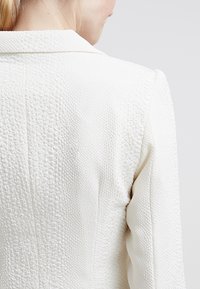 Blazer blanc texturé avec un col classique et une coupe ajustée, doté d'un tissu à motifs en relief et de coutures structurées à l'arrière.