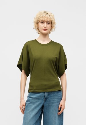 Donna con capelli biondi ricci che indossa una maglia verde oliva a maniche larghe e jeans larghi azzurri, in piedi davanti a uno sfondo neutro.