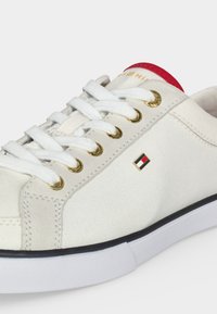 Λευκό καμβαδένιο αθλητικό παπούτσι με λευκά κορδόνια, χρυσά ρούμπελα, μικρό κόκκινο, λευκό και μπλε λογότυπο στο πλάι και κόκκινη λεπτομέρεια στον αστράγαλο.