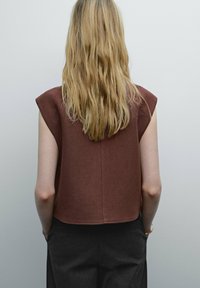 Avskuren, brun ärmlös topp med strukturerade axlar och subtil textur, kombinerad med mörkgrå byxor, sedd bakifrån.