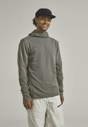 Smiling man wearing a dark gray textured hoodie with thumb holes and light beige cargo pants standing against a plain background.