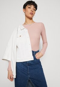 Rosa gerippter Langarmshirt, kombiniert mit einer weißen Jeansjacke, die über der Schulter getragen wird, und einem dunkelblauen Jeansrock mit einem Schlitz vorne.