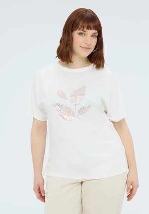 Kvinde med rødt hår iført hvid T-shirt med metallisk bladmotiv og beige bukser, stående mod en ensfarvet lys baggrund.