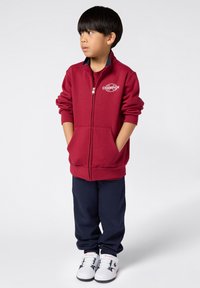 Bordeauxkleurige ritssluiting jack met logo, twee voorzakken, gecombineerd met marineblauwe broek en witte sneakers met zwarte accenten. Glad stoffen textuur.