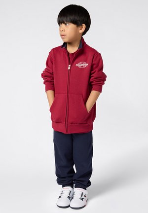 Bordeauxkleurige ritssluiting jack met logo, twee voorzakken, gecombineerd met marineblauwe broek en witte sneakers met zwarte accenten. Glad stoffen textuur.