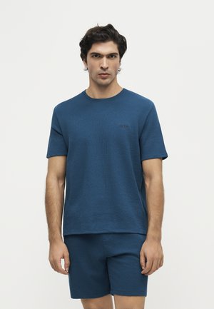 Homme portant une chemise à manches courtes bleue texturée assortie et un short, debout devant un fond blanc uni.