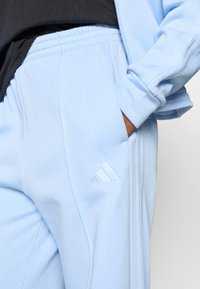 Osoba v svetlomodrých teplákových nohaviciach Adidas a zodpovedajúcej bunde, s rukou v vrecku, čierne tričko viditeľné na páse.